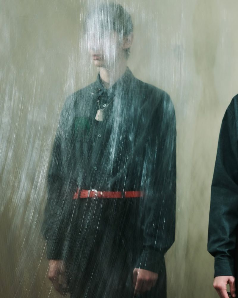 Two men in dark shirts with an orange belt stand partially obscured by falling water in a minimalist, monochromatic setting.