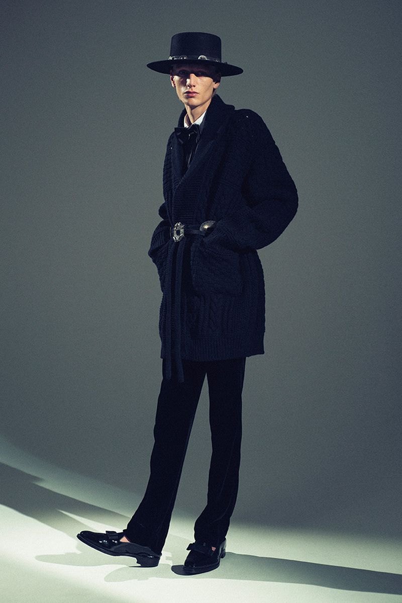 Full-length portrait of a young adult male fashion model wearing a black cowboy hat, knit cardigan, and trousers in a studio setting.