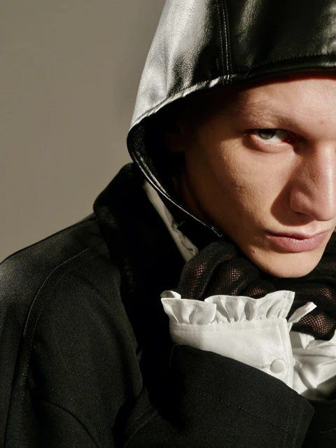 Close-up studio shot of a fair-skinned person in a black hooded garment with white ruffled sleeves. Dark jacket is visible.