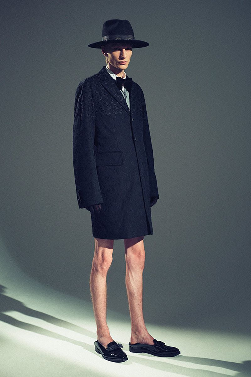 Full-length portrait of a slender male fashion model wearing a dark suit coat, bowler hat, and loafers against a neutral gray backdrop.