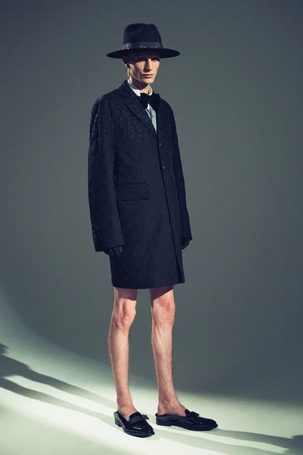 Full-length portrait of a slender male fashion model wearing a dark suit coat, bowler hat, and loafers against a neutral gray backdrop.