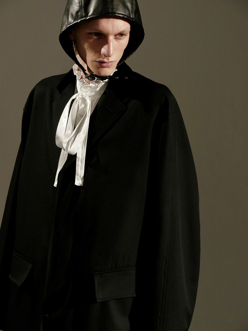 Fashion portrait of a person wearing a black jacket and a leather hat with a ruffled collar and white satin bow, studio lighting