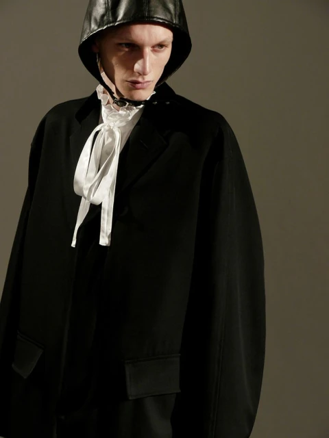 Fashion portrait of a person wearing a black jacket and a leather hat with a ruffled collar and white satin bow, studio lighting