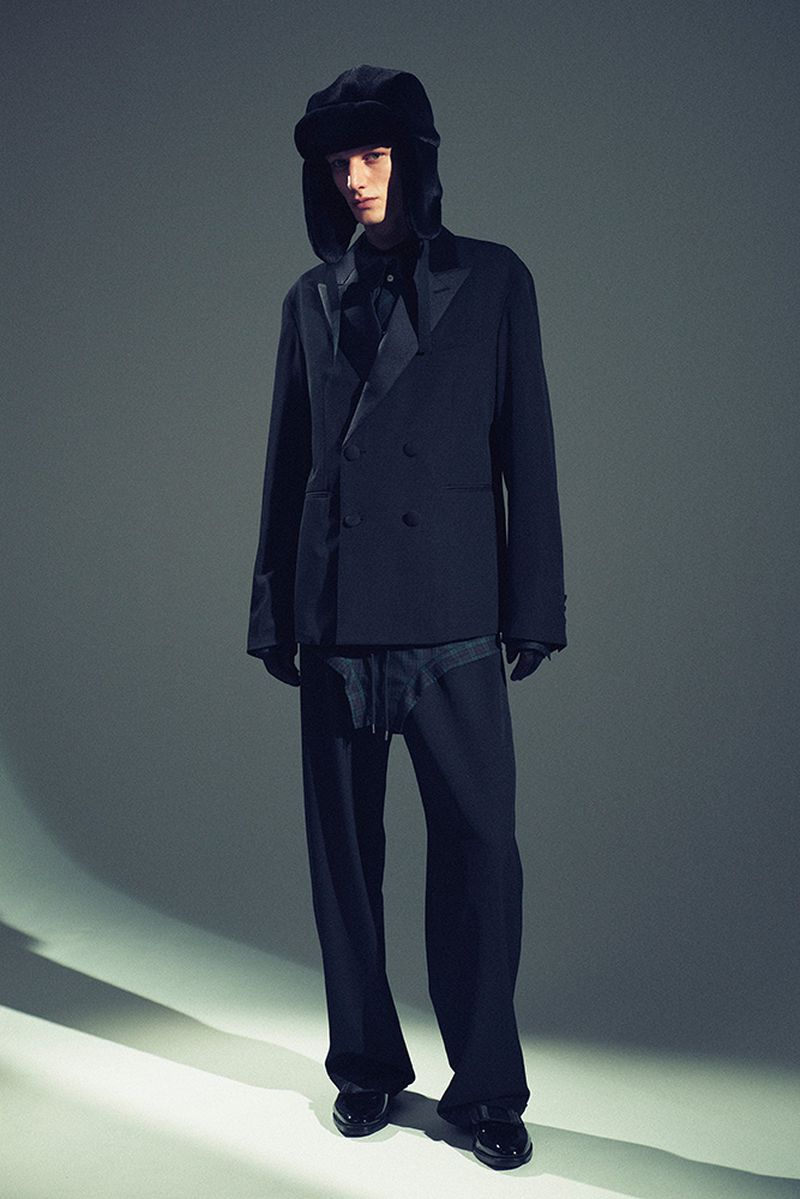 Man poses in black suit and furry hat in a fashion forward shot. Studio lighting highlights the textures and tailoring in a full shot.