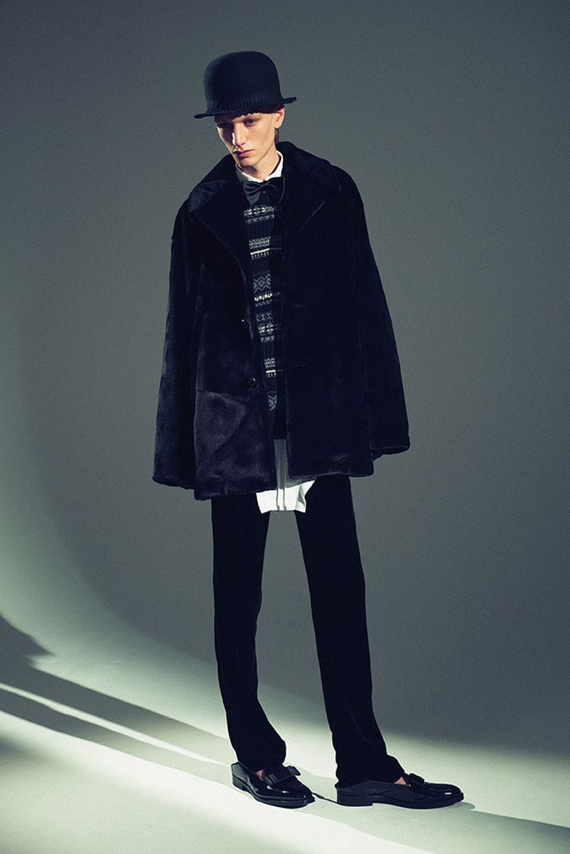 Full-length studio shot of a man wearing a bowler hat, fur coat, patterned sweater, trousers, and loafers, standing against a grey background.