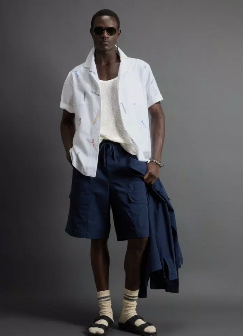 Man in light patterned shirt, white tank top, dark shorts, and sandals stands against a gray background, holding a jacket.
