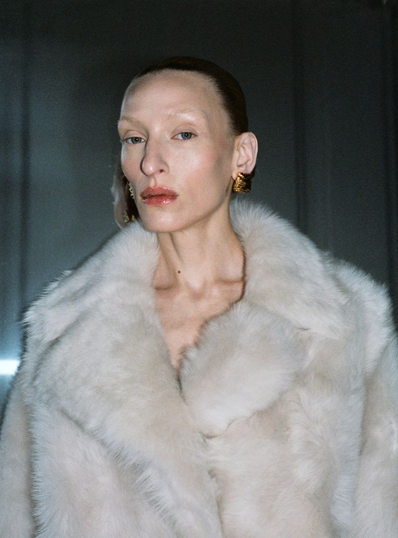 Headshot of an adult woman wearing a light faux fur coat and gold hoop earrings against a neutral backdrop in a high fashion setting.