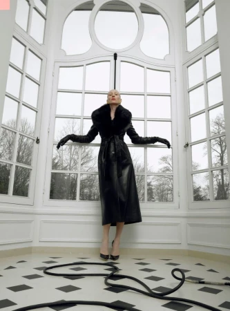 A striking fashion portrait of a woman in a black leather coat and long gloves, standing in a white-walled room with large windows and a black cable on the floor.