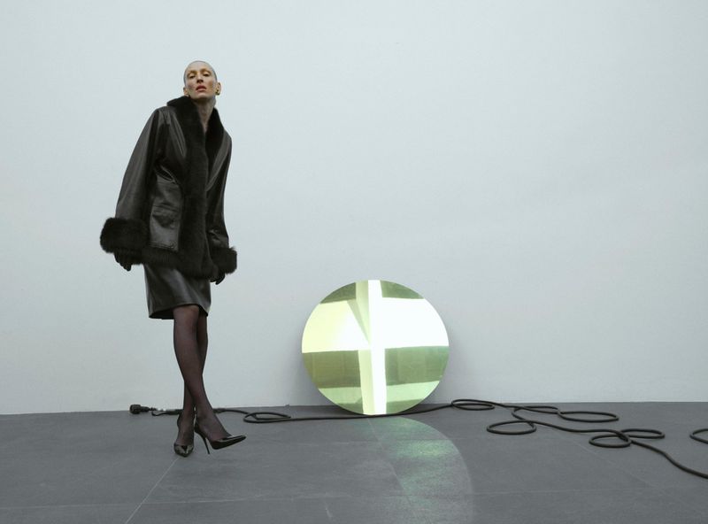 Woman in a leather coat and skirt standing near a circular light fixture in a photo studio with a white wall and gray floor.