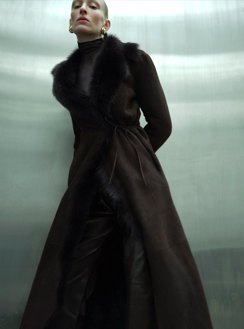 A slender woman with a shaved head poses in a long brown fur-trimmed coat against a metallic background, showcasing a fashion-forward look.