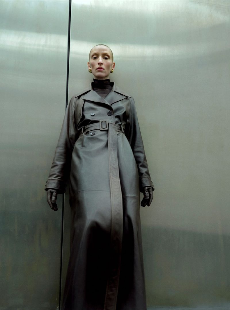 A bald woman with green earrings and a neutral expression wears a dark leather trench coat against a metallic wall.