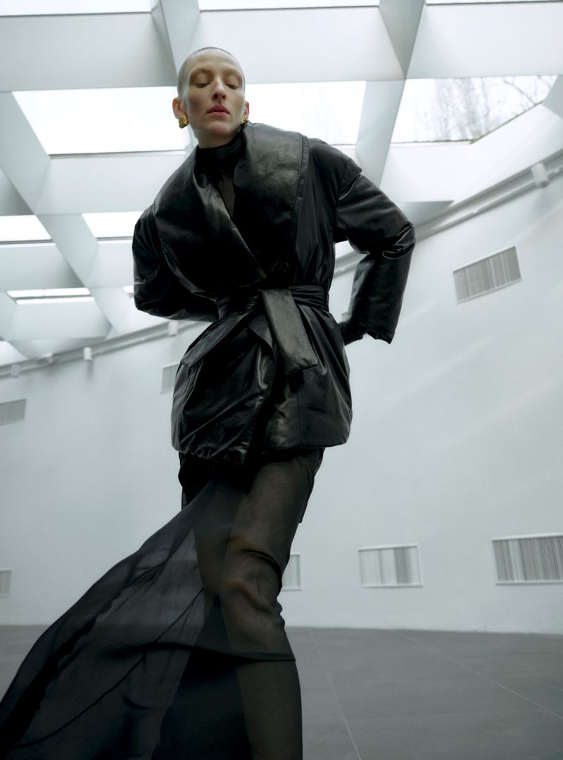 Fashion shot of a stylish woman in a black coat and long dress posing in a spacious, light-filled interior. High fashion look.