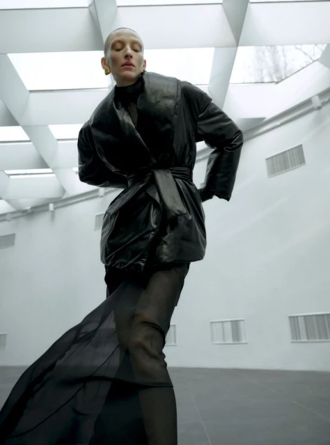 Fashion shot of a stylish woman in a black coat and long dress posing in a spacious, light-filled interior. High fashion look.