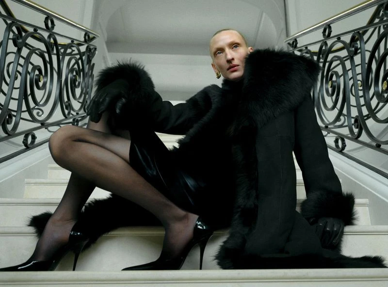 Woman in a black fur-trimmed coat and high heels poses on marble stairs with ornamental iron railing in an indoor setting.