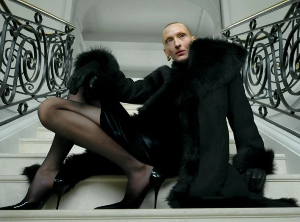 Woman in a black fur-trimmed coat and high heels poses on marble stairs with ornamental iron railing in an indoor setting.