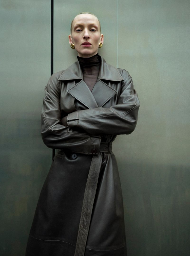 Adult female with slicked back hair and gold earrings poses in front of a grey metal panel wearing a dark brown leather trench coat.