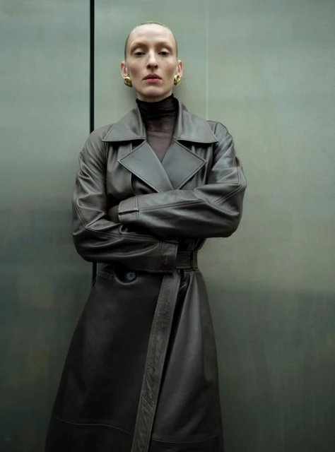 Adult female with slicked back hair and gold earrings poses in front of a grey metal panel wearing a dark brown leather trench coat.