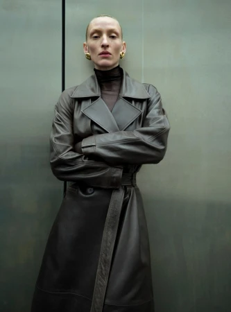 Adult female with slicked back hair and gold earrings poses in front of a grey metal panel wearing a dark brown leather trench coat.