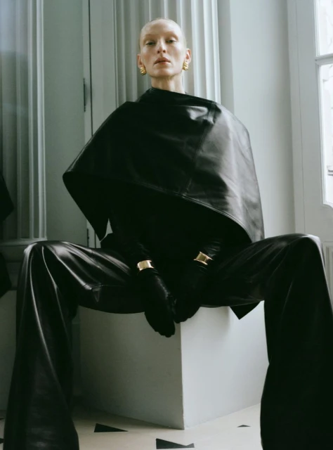 A bald woman poses in black leather-look attire. The setting is a minimalistic indoor studio with neutral tones.