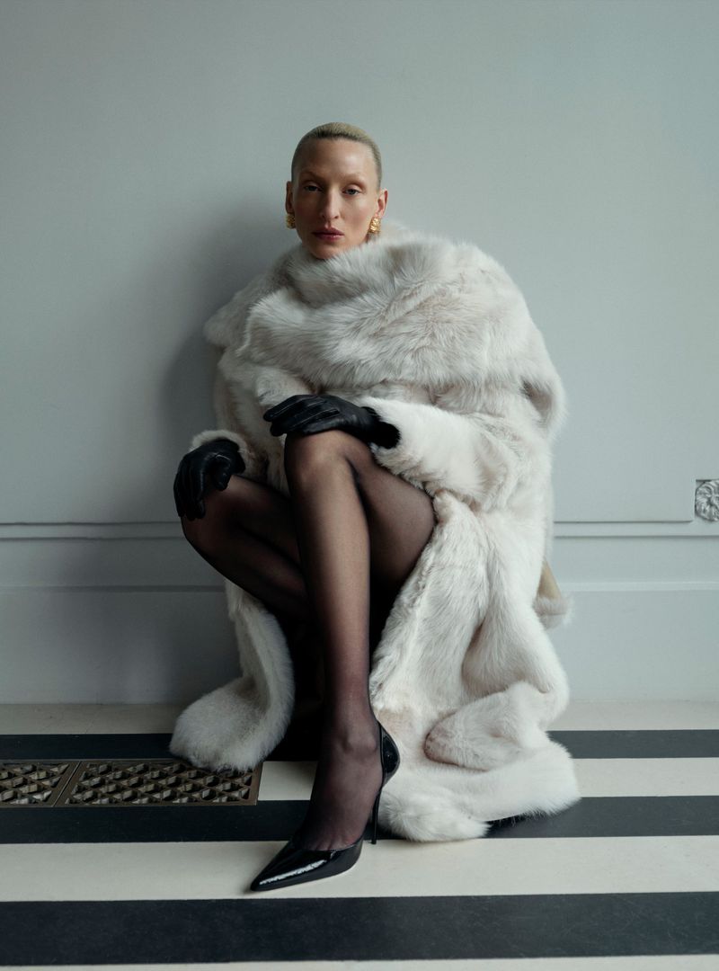 Adult female in fur coat, black gloves and high heels poses against a grey wall with striped floor in fashion studio setting.