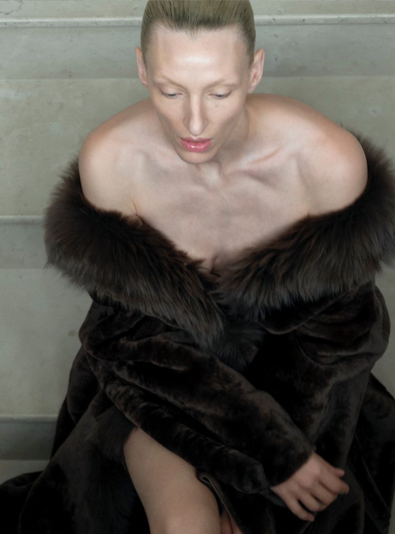 Fashion shot of an adult female in an off-the-shoulder brown fur coat sitting on steps with a neutral expression and fair skin.