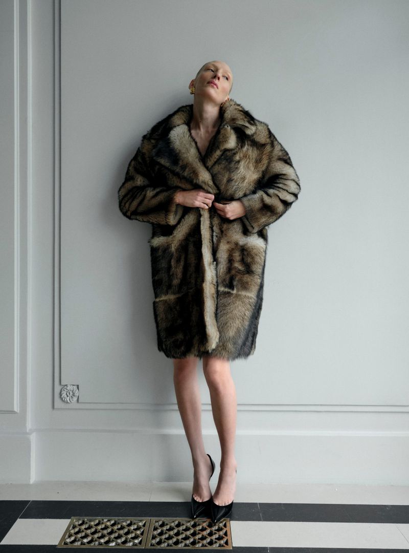 Woman in a fur coat standing against a wall, wearing black high heels, posing for a fashion shot in an indoor setting.