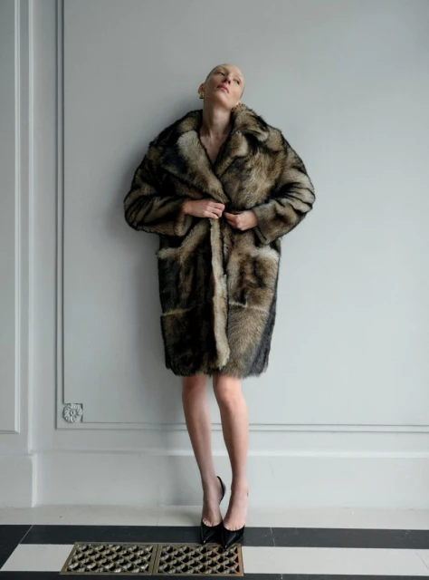 Woman in a fur coat standing against a wall, wearing black high heels, posing for a fashion shot in an indoor setting.