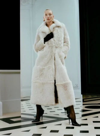 A woman poses in a full-length, cream-colored fur coat, black gloves, and black high heels in an indoor fashion shot with white walls.
