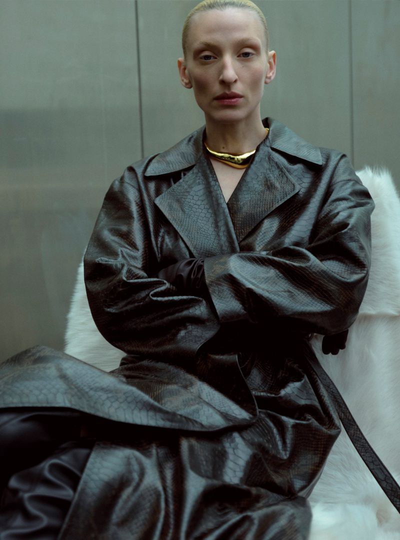 An adult female with a shaved head, wearing a reptile-print trench coat and gold necklace, sitting in an interior setting with white furry chair.