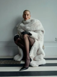Model wearing a voluminous white fur coat and black gloves crouching against a pale wall with striped flooring.
