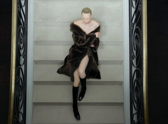 Model wearing a brown fur coat and black boots reclines on light stone steps between ornate iron railings.