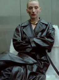 Model wearing a dark embossed leather trench coat with arms crossed, seated against a plain background