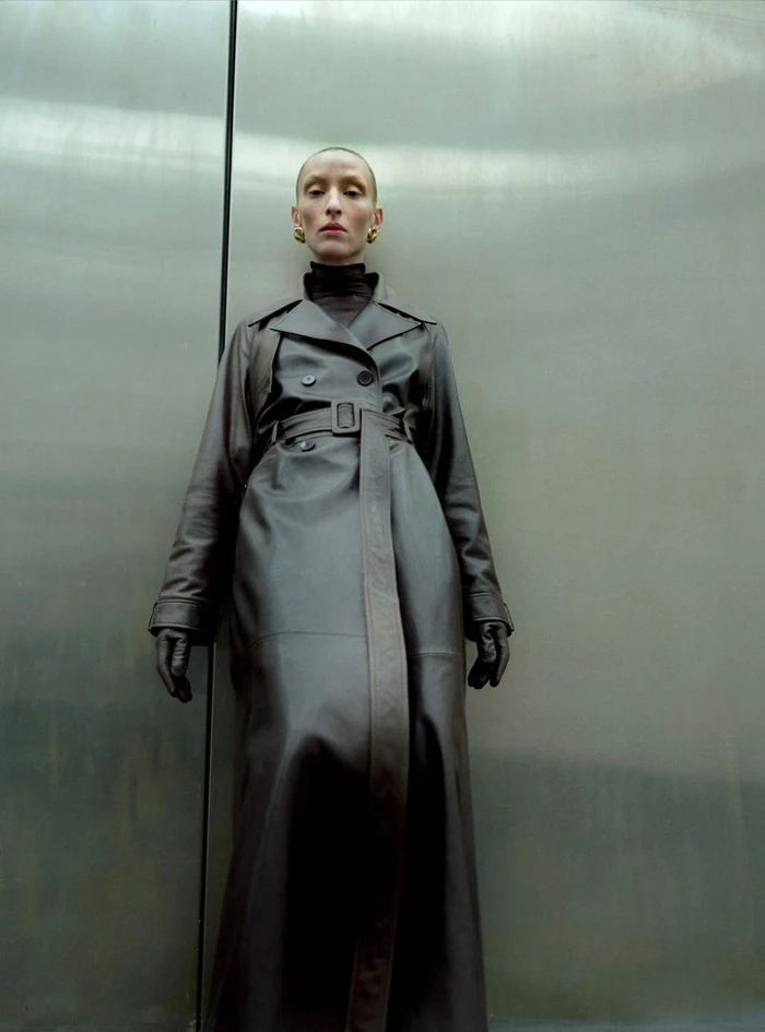 Model Maggie Maurer wearing a long brown leather trench coat against a sleek metal background for Nour Hammour.