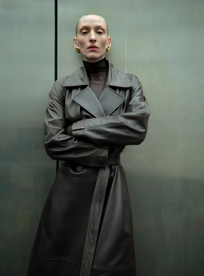 Model with shaved head wearing a dark brown leather trench coat against a textured green background