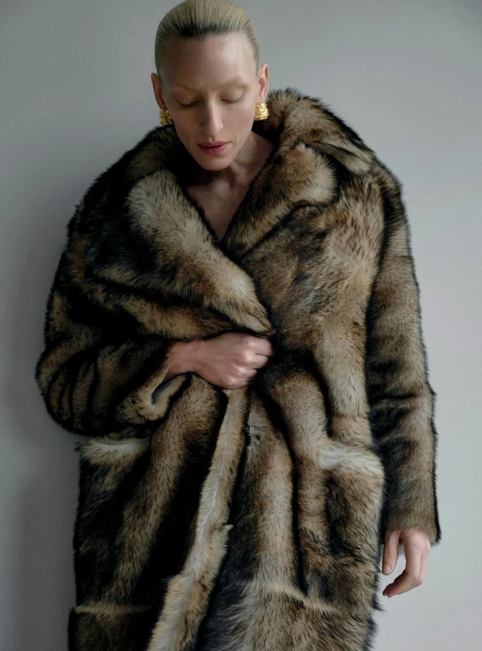 Model Maggie Maurer wearing a voluminous faux fur coat and gold earrings against a plain grey studio wall.