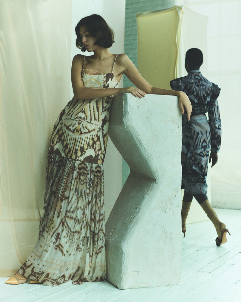 Two women in patterned dresses pose indoors with a geometric sculpture. One model stands in the foreground, the other fades into the background.