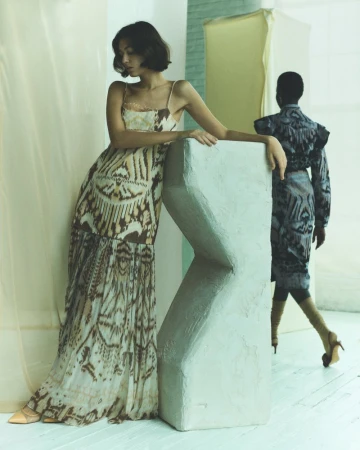 Two women in patterned dresses pose indoors with a geometric sculpture. One model stands in the foreground, the other fades into the background.