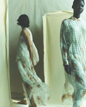 Two women model patterned dresses in a studio setting. One wears a long-sleeved dress with a belt, while the other sports a sleeveless dress.