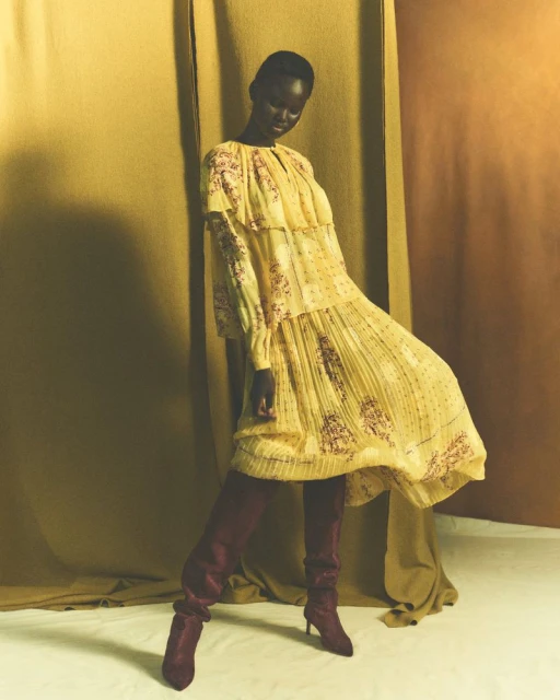 Fashion model in a flowing yellow floral dress and burgundy boots poses against a textured mustard background in a studio shot.
