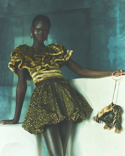 A strikingly beautiful Black female model poses in a golden ruffled top and patterned mini skirt against a muted blue background.