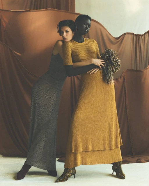 Two women pose in dresses against a draped brown fabric backdrop, one in gold and the other in dark gray, showcasing fashion and style.