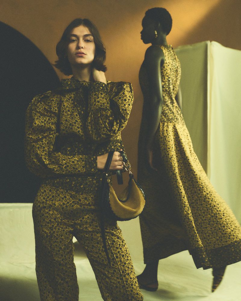 Two women pose in patterned outfits in a fashion editorial. One wears a jumpsuit and carries a handbag, the other wears a long dress.