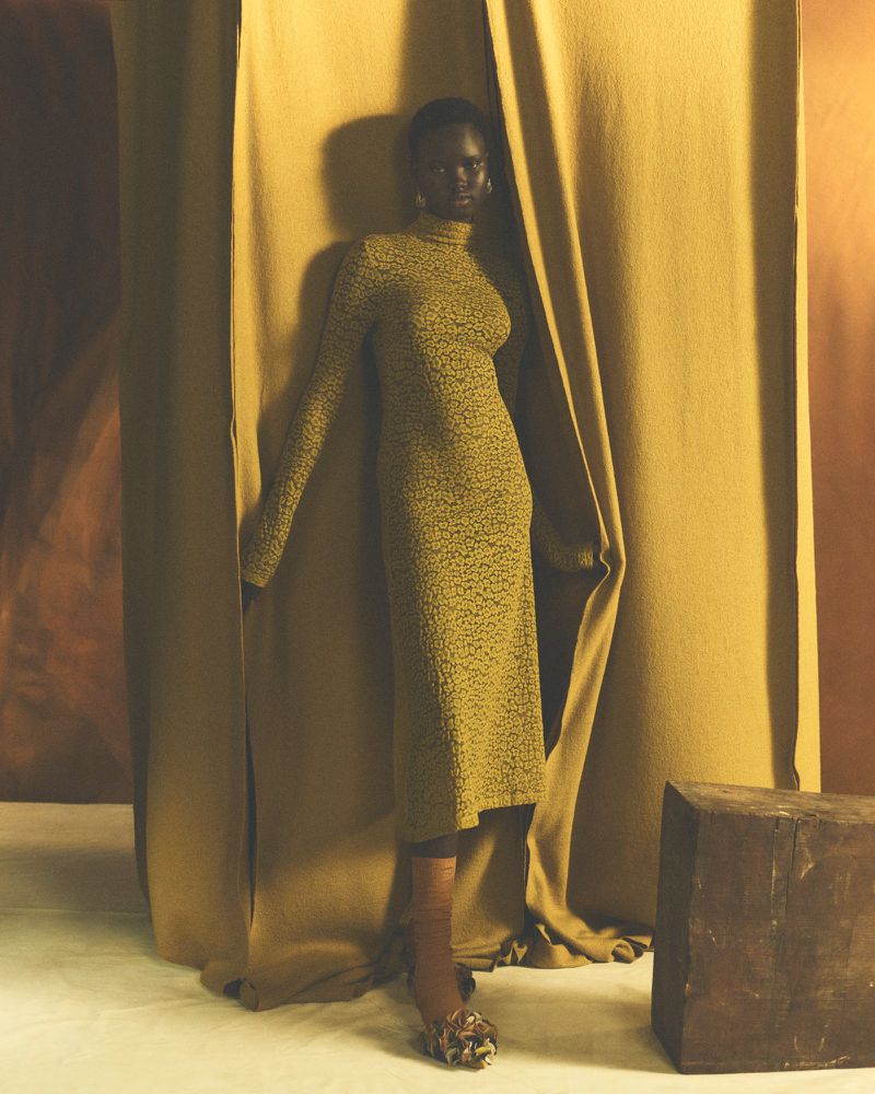 Elegant dark-skinned woman in patterned yellow dress poses confidently in a fashion-forward studio shot with draped fabric.