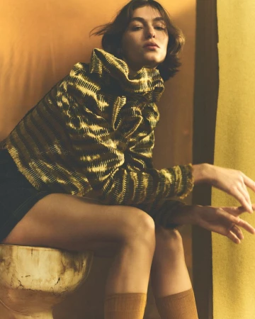 Brunette woman in a printed top and knee socks sits on a weathered, marble-like base in a golden-toned studio setting.