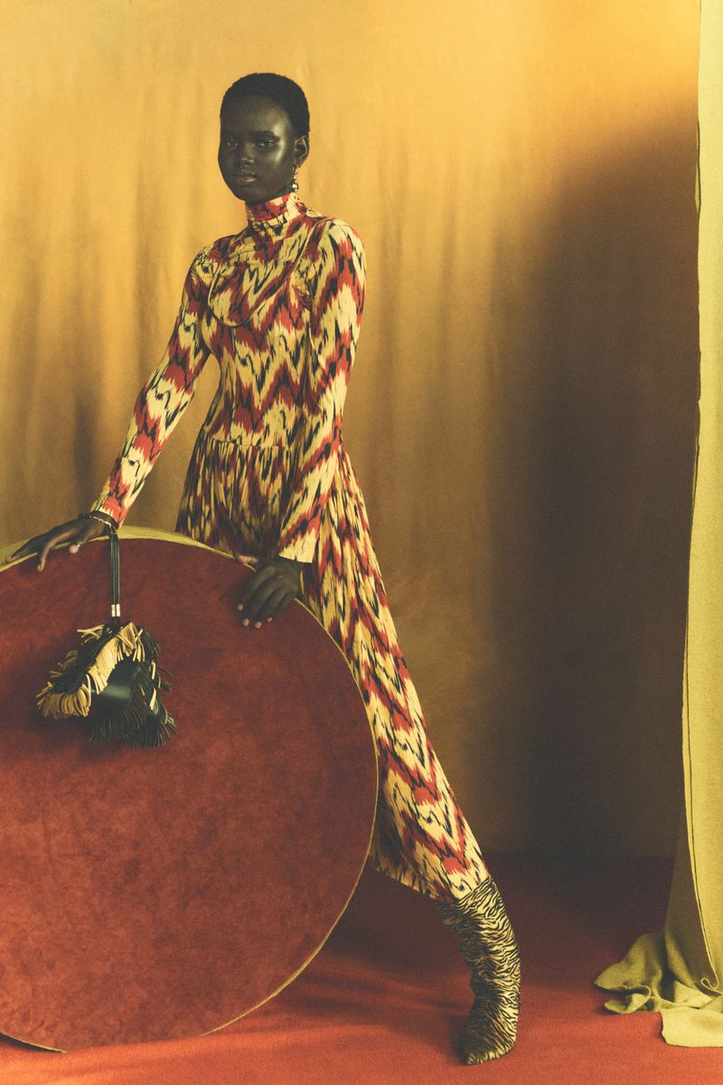 Fashion model in patterned jumpsuit poses with round table in a studio setting with textured background, showcasing style and design.