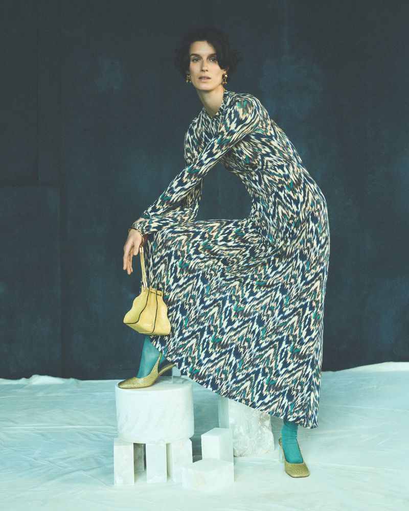 Full-length portrait of a female model wearing a patterned maxi dress, teal tights, and gold heels while posing indoors.