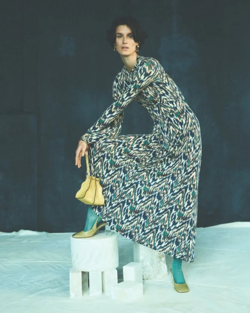 Full-length portrait of a female model wearing a patterned maxi dress, teal tights, and gold heels while posing indoors.