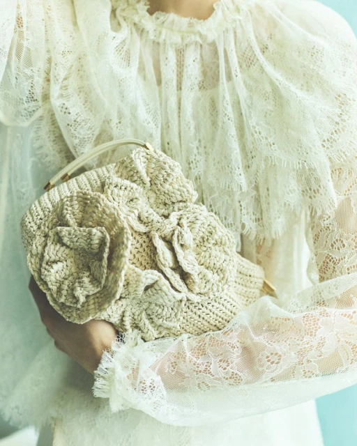 Close-up of a woman holding a decorative, textured handbag with a floral design, wearing a lace-trimmed blouse with ruffles.