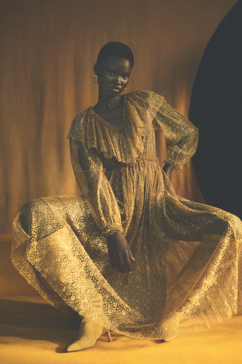 A dark-skinned woman in a sparkling gold gown poses in a seated position on a yellow surface in a dimly lit, indoor setting.
