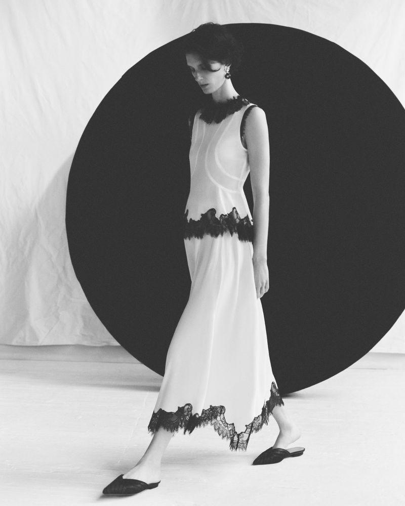 Monochrome fashion shot of an adult female model in a sleeveless white dress with black lace trim, walking against a black and white backdrop.